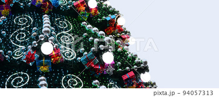 A fragment of a huge Christmas tree with many ornaments, gift boxes and luminous lamps. Photo of a decorated Christmas tree close-up with copy space 94057313