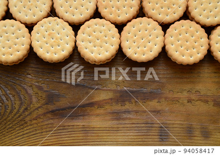 A round sandwich cookie with coconut filling lies in large quantities on a brown wooden surface. Photo of edible treats on a wooden background with copy space 94058417