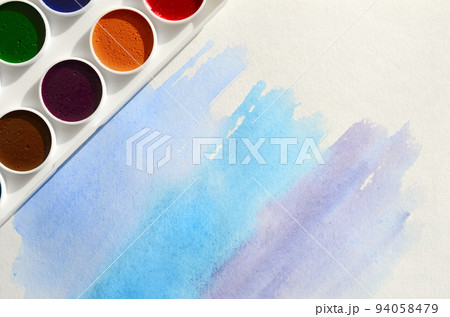 A new set of watercolors lies on a sheet of paper, which shows an abstract watercolor drawing in the form of blue strokes. The concept of amateur painting among teenagers A new set of watercolors lies on a sheet of paper, which shows an abstract watercolor drawing in the form of blue strokes. The concept of amateur painting among teenagers 94058479