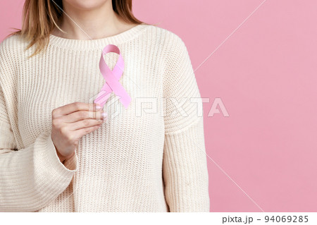 Female health check awareness. Closeup of woman hand in white pullover holding pink ribbon, symbol of breast cancer, timely diagnosis, support of oncology patients. indoor isolated on pink background 94069285