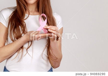 Female health check awareness. Closeup of woman hand in white pullover holding pink ribbon, symbol of breast cancer, timely diagnosis, support of oncology patients. indoor isolated on gray background Female health check awareness. Closeup of woman hand in white pullover holding pink ribbon, symbol of breast cancer, timely diagnosis, support of oncology patients. indoor isolated on gray background 94069290