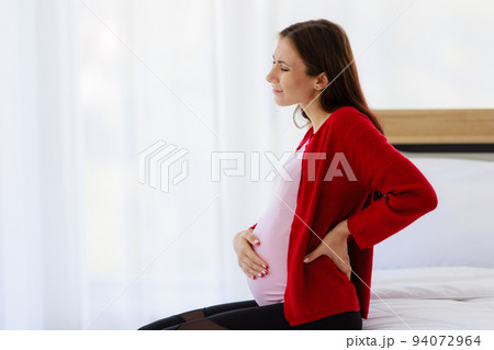 Pregnant woman sits on bed in bedroom, holding her back, showing pain or painful back. 94072964