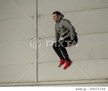 Caucasian man in hoodie jumping high with hands on knees outdoors.  94073148