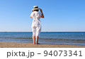 back view on a mature woman in a straw hat and white dress walks along the blue sea coast on a sunny 94073341