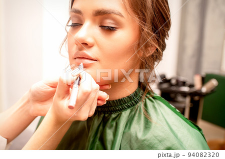 Lips makeup. Makeup artist applies contour with a pencil to the lips of a young woman in a beauty salon Lips makeup. Makeup artist applies contour with a pencil to the lips of a young woman in a beauty salon 94082320