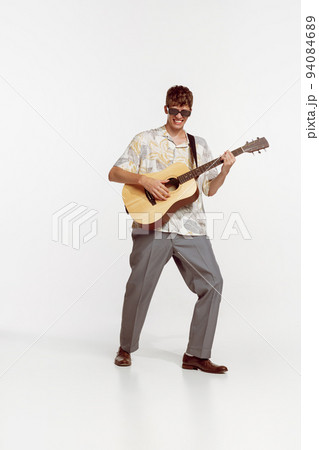 Portrait of young ma in sunglasses and vinatge clothes playing guitar, posing isolated over white studio background 94084689