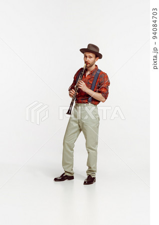 Portrait of stylish young man in red checkered shirt and hat playing clarinet isolated over white background. Romantic music 94084703