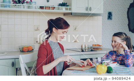 Cheerful happy mother cutting tomatoes while cooking and talking to her smiling daughter feeding mom in the kitchen at home. Family, cook, and people concept Cheerful happy mother cutting tomatoes while cooking and talking to her smiling daughter feeding mom in the kitchen at home. Family, cook, and people concept 94089424