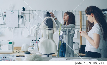 Young woman is measuring mannequin parts with tape-measure while her female colleague is entering this information in smartphone. Productive cooperation in tailoring business concept. 94089714