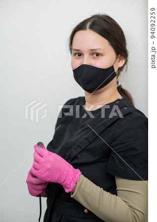 The pedicure master stands against the background of a white wall and holds an apparatus with an abrasive disc.  94092259