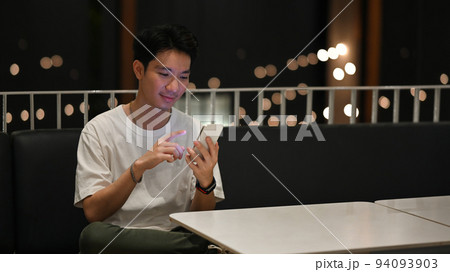 Young hipster man using smart phone while sitting at terrace with blurred night street lights background 94093903