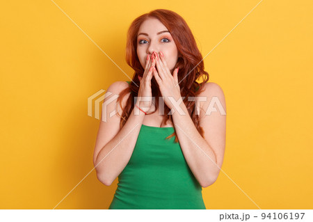 Beautiful young woman covers mouth with both palms, stares with shocking expression, wears green t shirt, has fashionable manicure, models over yellow background with surprise facial expression. Beautiful young woman covers mouth with both palms, stares with shocking expression, wears green t shirt, has fashionable manicure, models over yellow background with surprise facial expression. 94106197