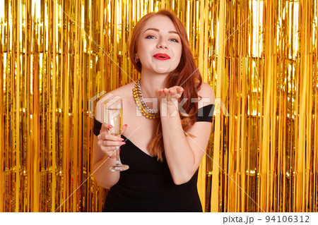Excited pretty red haired girl holding wineglass of sparkling champagne standing isolated on sparkle background. Model sending air kisses while celebrating important event. Excited pretty red haired girl holding wineglass of sparkling champagne standing isolated on sparkle background. Model sending air kisses while celebrating important event. 94106312