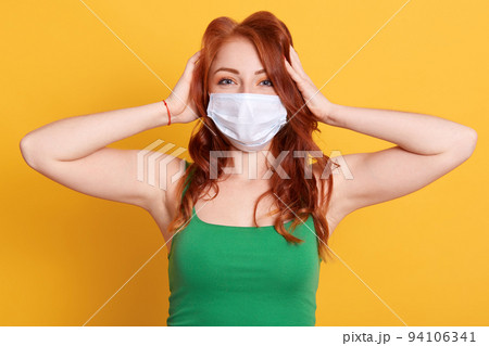 Woman in medical mask plugging ears with hands. European young redhead female ignoring noise or news about coronavirus, wearing green t shirt, posing against yellow wall. Woman in medical mask plugging ears with hands. European young redhead female ignoring noise or news about coronavirus, wearing green t shirt, posing against yellow wall. 94106341
