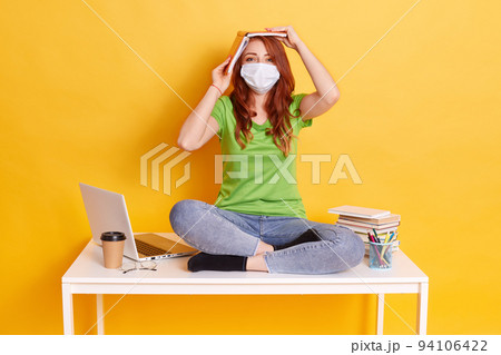 Red haired girl in medical mask sits with crossed legs on table, wearing jeans and green t shirt, being tired of long ours distance learning, poses against yellow wall. 94106422