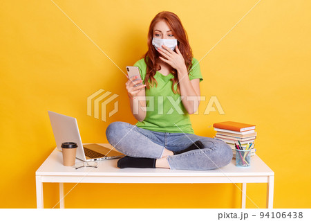 Surprised young woman got news via text message while learning, holding mobile phone and covering her mouth with palm, wear medical mask and casual clothing, sitting on table with crossed legs. Surprised young woman got news via text message while learning, holding mobile phone and covering her mouth with palm, wear medical mask and casual clothing, sitting on table with crossed legs. 94106438