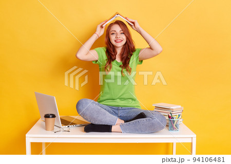 Young attractive girl with book over her head, like symbol of roof or house, looks smiling directly at camera, wearing casual closing, having fun while preparing for classes, against yellow background Young attractive girl with book over her head, like symbol of roof or house, looks smiling directly at camera, wearing casual closing, having fun while preparing for classes, against yellow background 94106481