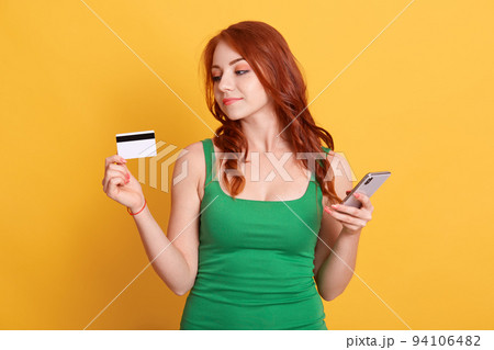 Portrait of nice attractive red haired girl holding in hands bank card and cell phone, wireless purchase, ginger girl buying things online isolated over yellow background. Portrait of nice attractive red haired girl holding in hands bank card and cell phone, wireless purchase, ginger girl buying things online isolated over yellow background. 94106482
