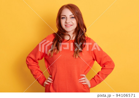 Confident young lady with red hair standing with hands on hips and looking at camera, being in good mood, smiling happily against yellow background, female wearing casual attire. Confident young lady with red hair standing with hands on hips and looking at camera, being in good mood, smiling happily against yellow background, female wearing casual attire. 94106571
