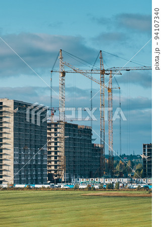 Building site background. Industrial construction cranes on background of clouds sky. Hoisting cranes, multi-storey buildings of new city districts and large green field. Project of urban area. 94107340