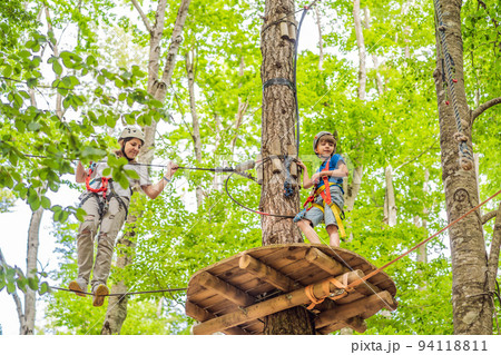 Mother and son climbing in extreme road trolley zipline in forest on carabiner safety link on tree to tree top rope adventure park. Family weekend children kids activities concept Portrait of a Mother and son climbing in extreme road trolley zipline in forest on carabiner safety link on tree to tree top rope adventure park. Family weekend children kids activities concept Portrait of a 94118811
