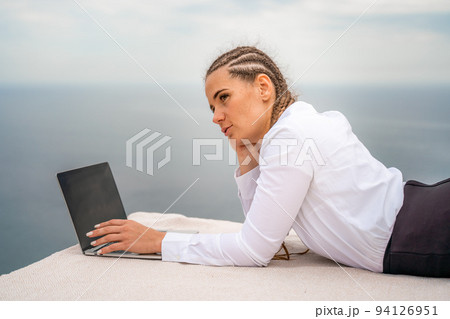 A woman is lying and typing on a laptop keyboard on a terrace with a beautiful sea view. Wearing a white blouse and black skirt. Freelance travel and vacation concept, digital nomad. A woman is lying and typing on a laptop keyboard on a terrace with a beautiful sea view. Wearing a white blouse and black skirt. Freelance travel and vacation concept, digital nomad. 94126951