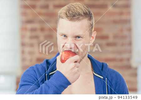 Close up photo. Portrait of young handsome athletic athletic man eating apple in kitchen at home, making fresh juice and fruit salad, looking at camera 94133549