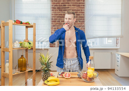 Young handsome athletic man eating apple in kitchen at home, making fresh juice and fruit salad, looking at camera Young handsome athletic man eating apple in kitchen at home, making fresh juice and fruit salad, looking at camera 94133554