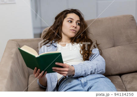 Young beautiful woman lying at home on the couch and reading a book, resting, looking at the camera, smiling Young beautiful woman lying at home on the couch and reading a book, resting, looking at the camera, smiling 94134250