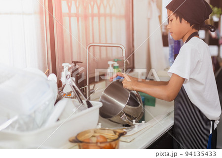 Young boy with apron doing the dishes at kitchen sink in the house. Young boy with apron doing the dishes at kitchen sink in the house. 94135433