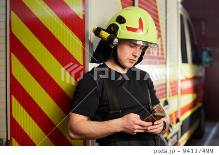 Portrait of a firefighter standing in front of a fire engine. Fireman using smart phone 94137499
