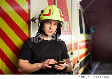 Portrait of a firefighter standing in front of a fire engine. Fireman using smart phone Portrait of a firefighter standing in front of a fire engine. Fireman using smart phone 94137500