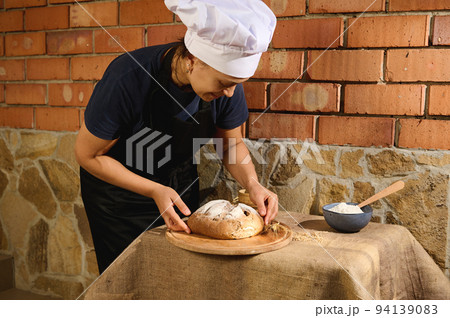Female baker in chef's cap and black apron, puts a homemade sourdough bread on a wooden board in artisanal family bakery Female baker in chef's cap and black apron, puts a homemade sourdough bread on a wooden board in artisanal family bakery 94139083