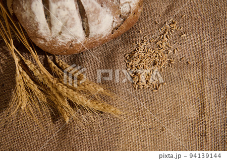 Still life. Wheat ears, partial view of a homemade whole grain bread and cereal grains scattered on a burlap tablecloth 94139144