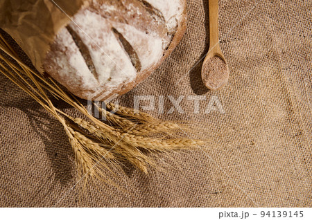 A loaf of whole grain bread sprinkled with flour, spikelets of wheat and a wooden spoon with bran on sacklock tablecloth A loaf of whole grain bread sprinkled with flour, spikelets of wheat and a wooden spoon with bran on sacklock tablecloth 94139145