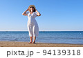 mature woman in sunglasses in a straw hat and white dress walks along the blue sea coast on a sunny 94139318