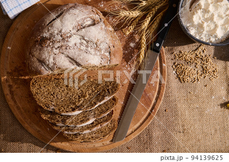 Top view fresh bread with crumbs on wooden board, spikelets of wheat and scattered cereal grains on a burlap tablecloth 94139625
