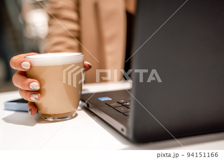 Close-up of beautiful female hands holding a large white cup of cappuccino. A business woman sits in a cafe working at a computer. 94151166