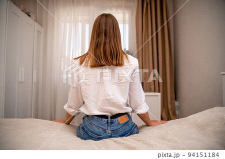 A young woman in a white shirt and jeans sits on the bed with her back against the background of the room and the window. Home furnishings. 94151184