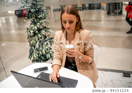 A business woman sits in a cafe, works at a computer, drinks coffee. She is wearing a beige jacket, brown top and black trousers. 94151224