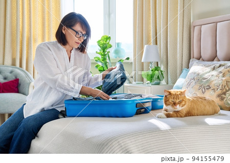 Middle-aged woman packing suitcase, sad cat lying on the bed Middle-aged woman packing suitcase, sad cat lying on the bed 94155479