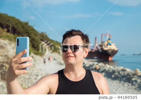 Happy smiling man taking selfie with phone outdoors against sunken cargo ship. 32 years old Caucasian tourist man. Close-up summer headshot lifestyle three quarter portrait. Travel, Vacation, portrait Happy smiling man taking selfie with phone outdoors against sunken cargo ship. 32 years old Caucasian tourist man. Close-up summer headshot lifestyle three quarter portrait. Travel, Vacation, portrait 94160505