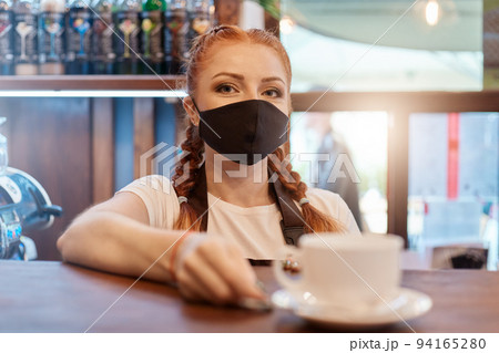 Red haired young barista girl in medical mask with city cafe background, offers cup of coffee to client, bartender near counter, safety during pandemic. 94165280