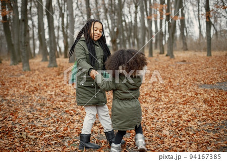 Beautiful little black girls standing near tent in the forest. Two little sisters dancing in autumn forest. Black girls wearing khaki coats. 94167385