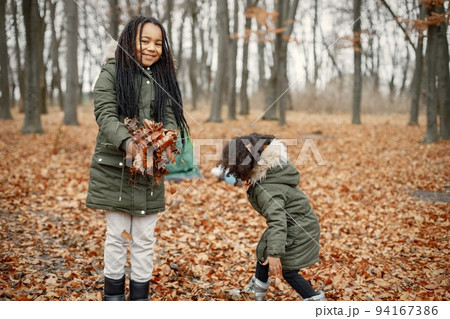 Beautiful little black girls standing near tent in the forest. Two little sisters playing with yellow leaves in autumn forest. Black girls wearing khaki coats. 94167386