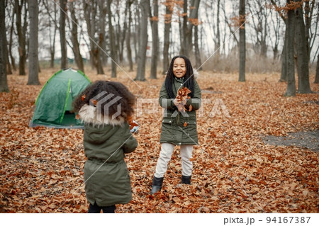 Beautiful little black girls standing near tent in the forest. Two little sisters playing with yellow leaves in autumn forest. Black girls wearing khaki coats. 94167387
