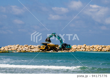 An excavator and a mining truck move stones into the sea. Construction of breakwaters to protect the beach in Netanya in Israel. 94175776