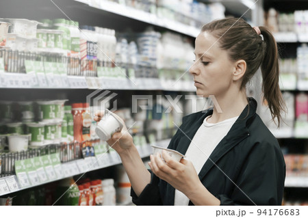 a young lady in the store chooses yogurt products, reads and compares the composition on the labels. The concept of healthy eating low-fat foods a young lady in the store chooses yogurt products, reads and compares the composition on the labels. The concept of healthy eating low-fat foods 94176683