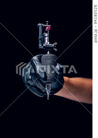 Machine for tattoo. Human hand in black protective glove holding professional equipment for making tattoo on body isolated on dark background. Machine for tattoo. Human hand in black protective glove holding professional equipment for making tattoo on body isolated on dark background. 94180526