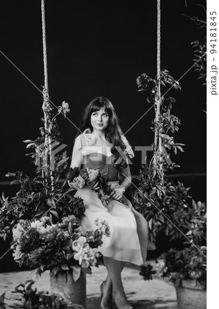 an attractive woman in a dress sits on a swing in peony flowers. black and white an attractive woman in a dress sits on a swing in peony flowers. black and white 94181845